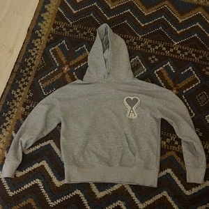 Grå hoodie från AMI Paris med hjärtdetalj - Säljer en grå hoodie från AMI Paris med deras ikoniska hjärta-A patch på bröstet. Tröjan har huva, känguruficka och är tillverkad i mjuk bomull. Perfekt för chill dagar och skön avslappnad stil. Sitter S-M