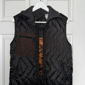 Svart quiltad dunväst från Error - Svart quiltad dunväst från Error med grå ribbad krage och brunt bröstficka med dragkedja. Snyggt mönstrad insida med orange tryck och flera dragkedjefickor framtill. Perfekt för lager-på-lager och stilren look!