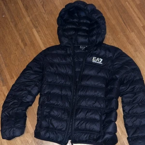 Svart dunjacka EA7 Emporio Armani - Säljer en svart dunjacka från EA7 Emporio Armani med huva och dragkedja. Jackan har quiltad design och logga på bröstet. Perfekt för kyliga dagar och har en snygg, sportig look. Passformen är normal och jackan är lätt men ändå varm.  OBS: Jackan har en litet rivning nära skulderbladet som syns endast vid nära inspektion med ficklampa (bild).   Nypris: 1800, vid snabb affär kan priset prutas. Fråga gärna frågor vid köp. 