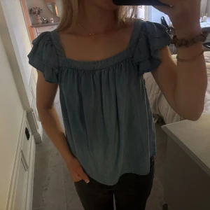 Denim volangblus med kort ärm - Söt denim blus med volangdetaljer på axlarna och korta ärmar. Blusen har en fin passform och är tillverkad i ett mjukt, jeansliknande material. Perfekt för en avslappnad och trendig look.