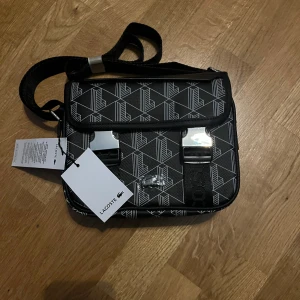 Lacoste Väska - Snygg svart helt oanvänd axelväska från Lacoste med geometriskt mönster i vitt. Väskan har justerbar axelrem, två silvriga spännen framtill och detaljer i syntetmaterial. Perfekt storlek för det viktigaste och har cool urban vibe med Lacoste-logga på framsidan.