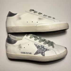 Golden Goose Superstar sneakers silver - Golden Goose Superstar sneakers i vitt skinn med silverfärgad häl och grå snörning. Sidan pryds av en glittrig silverstjärna och GGDB/SSTAR-tryck. Skorna har en vintage-inspirerad look med slitna detaljer och platt sula. UTFÖRSÄLJNING GÄLLER TILL 10 OKTOBER !!!