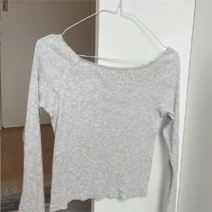 Ljusgrå ribbad offshoulder-top - Säljer en ljusgrå ribbad topp med offshoulder-skärning och långa ärmar. Toppen har en tight passform och är tillverkad i mjuk bomullsmix, perfekt för en clean och stilren look. Enkel att matcha med jeans eller kjol.