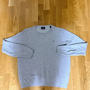 Ljusgrå stickad tröja från GANT - Snygg ljusgrå stickad tröja från GANT med rund hals och diskret logga broderad på bröstet. Tröjan har långa ärmar och ribbade muddar vid ärmslut och nederkant. Perfekt för dig som gillar klassisk och stilren look.