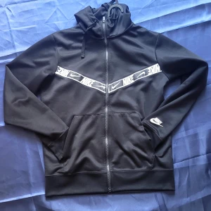 Svart hoodie från Nike med dragkedja - Svart hoodie från Nike i storlek S med vit logga på ärmen och coolt band med Nike-tryck över bröstet. Tröjan har huva med snörning, praktiska fickor och hel dragkedja framtill. Perfekt för chill eller träning.