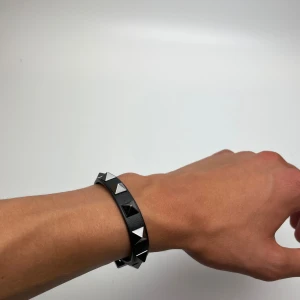 Valentino svart armband med nitar - Snyggt svart armband från Valentino Garavani i läder med silverfärgade pyramidformade nitar runt om. Justerbart spänne i metall och klassisk edgy look. Perfekt accessoar för att lyfta din outfit med lite attityd.