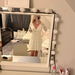 Vit spetsklänning med v-ringning - Superfin vit kort klänning i spets med långärmade ärmar och v-ringad fram. Klänningen har en markerad midja och lätt utställd kjol. Baksidan har fyrkantig urringning. Perfekt för dig som gillar romantisk och feminin stil.