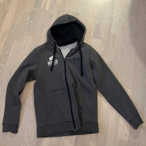 Nocco hoodie  - Mörkgrå hoodie från NOCCO i samarbete med Under Armour. Tröjan har dragkedja, huva med snörning och stora vita tryck både fram och bak. Perfekt för dig som gillar sportig stil och vill sticka ut på gymmet.