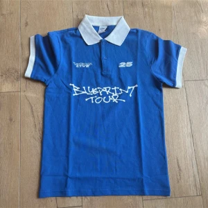 Blå pikétröja Rocboyz Blueprint Tour - Blå pikétröja från Rocboyz Studios med vit krage och ärmslut. Tryck på bröstet med 'Blueprint Tour' och siffran 25. Tillverkad i 100% bomull, klassisk passform och kortärmad. Perfekt för dig som gillar streetwear och snygga detaljer.