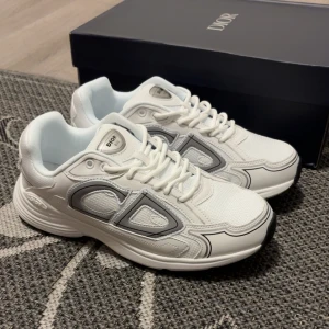 Dior B30 vita sneakers - Snygga Dior B30 sneakers i vitt med gråa detaljer och tydlig CD-logga på sidan. Skorna har en chunky sula i svart och vitt, mesh och skinnpaneler samt snörning. Perfekta för dig som gillar lyxiga och trendiga sneakers med sportig vibe.