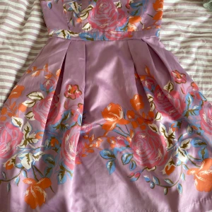 Ljuslila blommig klänning - Superfin ljuslila klänning med färgglada blommor i rosa, orange och blått. Klänningen har en utsvängd kjol med veck och en markerad midja. Perfekt för dig som gillar romantiska och färgstarka plagg.