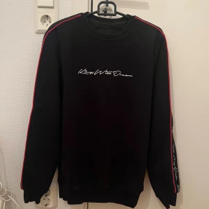 Svart Kings Will Dream sweatshirt XS - Svart sweatshirt från Kings Will Dream i storlek XS. Tröjan har röda ränder längs ärmarna och vita logotryck på bröstet och längs ärmarna. Perfekt för en streetwear-look.