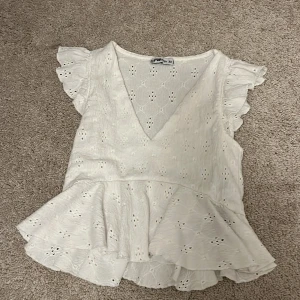 Vit volangblus från FB Sister XS - Supersöt vit blus från FB Sister i storlek XS. Blusen har broderade hålmönster, volangdetaljer vid ärmar och nederkant samt en djup v-ringning. Perfekt för sommarens alla tillfällen och ger en romantisk vibe. Lite kort på magen tycker jag som är 157 cm lång och storlek xs/xxs