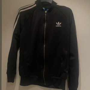Svart Adidas bomberjacka med vita ränder - Säljer en svart bomberjacka från Adidas med klassiska vita ränder längs ärmarna och Adidas-logga på bröstet. Jackan har dragkedja framtill, ribbade muddar och är långärmad. Perfekt för en sportig och stilren look.