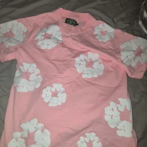 Rosa blommig t-shirt från Denim Tears s/M - Säljer en rosa t-shirt från Denim Tears med stora vita blommor tryckta över hela plagget. T-shirten är i mjuk bomull och har klassisk passform med korta ärmar. Perfekt för dig som vill sticka ut med en färgglad och unik stil.