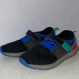 Svarta sneakers med blå detaljer - Snygga svarta sneakers med blå snören och blå sula. Skorna har en stickad ovandel, kardborreband med grönt spänne och röda detaljer på hälen. Perfekta för dig som gillar färg och sportig stil. Lätta och bekväma med platt sula. Eftersom den är gammal så står inte storleken på, men jag gissar på att den är 21