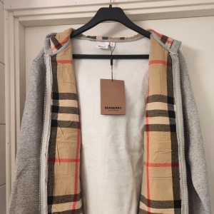 Grå hoodie från Burberry med dragkedja - Snygg grå hoodie från Burberry i storlek L. Tröjan har dragkedja, huva med snörning och klassiskt rutigt foder i luvan. Broderad Burberry-logga på bröstet och två fickor framtill. Perfekt för en chill och stilren look.