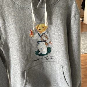 Grå hoodie Polo Ralph Lauren M - Grå hoodie från Polo Ralph Lauren med ikoniska Polo Bear-trycket på bröstet. Tröjan har huva med vita snören och en stor magficka. Mjuk insida och klassisk passform, perfekt för en chill och sportig stil.