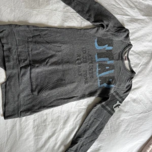 Grå långärmad hoodie från Replay - Mysig grå klänning från Replay med trycket 'STAR' i blått och svart framtill. Klänningen har magficka, långa ärmar och mjuk insida. Snygga detaljer på ena ärmen och avslappnad passform, perfekt för chill dagar.