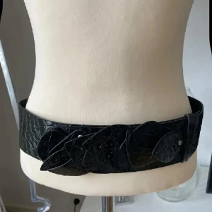 Brett svart bälte i skinn med bladdekor - Snyggt brett svart bälte i skinn med krokopräglad yta och dekorativa bladformade detaljer framtill. Bältet har en unik design som sticker ut och passar perfekt för att ge din outfit en cool touch. Ingen synlig logga eller märke.