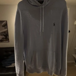 Ljusblå hoodie från Polo Ralph Lauren - Snygg ljusblå hoodie från Polo Ralph Lauren med klassisk broderad logga på bröstet. Tröjan är stickad i bomull, har huva med snörning och en stor magficka. Perfekt för dig som gillar en clean och stilren look.