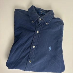 Blå skjorta från Ralph Lauren M - Snygg blå skjorta från Ralph Lauren i modellen Featherweight Mesh. Skjortan har klassisk krage, långa ärmar och vita knappar framtill. På bröstet finns den ikoniska broderade polospelaren i ljusblått. Perfekt för dig som gillar stilrena och tidlösa plagg.