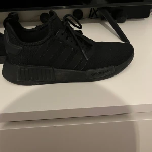 Svarta Adidas NMD sneakers - Stilrena helsvarta Adidas NMD sneakers med klassisk mesh-ovandel och snörning. Skon har en chunky sula och diskreta ränder på sidan. Perfekta för dig som gillar en clean och sportig look.