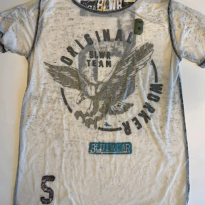 Vit t-shirt med tryck från Bluewear - Vit t-shirt från Bluewear med grått och svart tryck av örn och texten 'ORIGINAL WORKER BLWR TEAM'. Snygga kontrastsömmar, stort siffratryck på ryggen och detaljer i blått. Skön passform och cool streetstil. Genomskinlig 