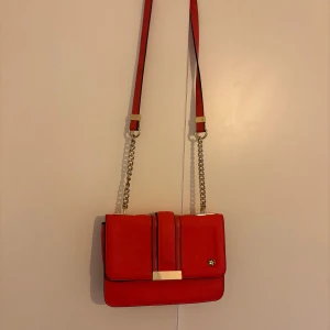 Röd axelväska med kedjerem - Snygg röd axelväska med guldfärgade kedjedetaljer och magnetlås. Väskan har ett blommigt innerfoder i svart och vitt samt praktiska innerfack med dragkedja. Stilren design med gulddetaljer framtill och justerbar rem i rött och guld. !Dock har den två fläckar på baksidan som inte går bort!