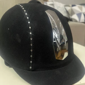 Svart ridhjälm från CRW med bling - Två ridhjälmar 130kr st.  En svart ridhjälm från CRW Classic Riding Wear.  Säljer även en café ridhjälm har sammetsliknande yta, snygg silverdetalj framtill och en rad med små glittrande stenar längs ovansidan. Perfekt för dig som vill sticka ut i stallet. Båda är i jättebra skick utan några defekter 
