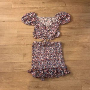 Färgglad kortkjol med volang från FB Sister - Superfin kortkjol och topp från FB Sister i smockad bomull med volangkant. Kjolen har ett färgglatt blommigt mönster i blått, rosa, orange och vitt. Perfekt för dig som gillar en lekfull och somrig stil.