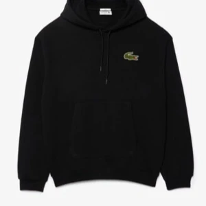 Svart zip från Lacoste med huva - Svart hoodie från Lacoste med klassisk krokodillogga på bröstet. Tröjan har huva med snörning och en stor magficka framtill. Perfekt för dig som gillar en enkel och stilren look.
