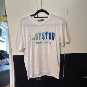 Vit Trapstar t-shirt med blått tryck - Säljer en clean vit t-shirt från Trapstar med stort broderat Trapstar-tryck i blå och silver/vit nyanser på bröstet. Klassisk passform med rund hals och korta ärmar. Tillverkad i mjuk bomull för skön känsla hela dagen. Aldrig använd nyskick.