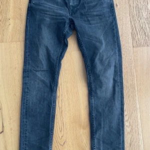 Svarta jeans från Nudie Jeans, W31 L32 - Säljer ett par svarta jeans från Nudie Jeans med klassisk femficksdesign och snygga detaljer på bakfickorna. Jeansen har en rak passform och är tillverkade i bomull. Perfekta för dig som gillar en stilren och avslappnad look.
