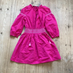 Rosa kort klänning från Zara - Snygg rosa klänning från Zara med smock i midjan och på ärmarna. Modellen har trekvartsärmar, volang vid halsen och knytband med tofsar.
