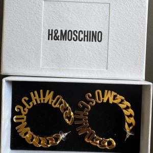 Guldiga örhängen H&M x Moschino - Säljer ett par statement örhängen från H&M x Moschino i guldigt metallmaterial. De är stora hoops med chunky kedjedetaljer och bokstäver som bildar 'MOSCHINO' runt hela ringen. Perfekta för dig som gillar att sticka ut med accessoarer.