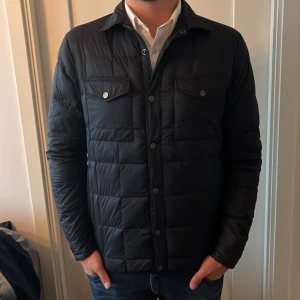 Mörkblå field jacket från East West - Svart quiltad jacka från East West med klassisk krage, tryckknappar och två bröstfickor med lock. Jackan har lång ärm och en stilren, rutig quiltning. Perfekt för dig som vill ha en clean och tidlös look.