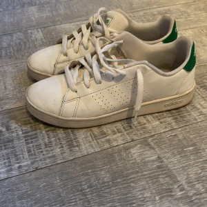 Adidas Stan Smith vita sneakers - Klassiska Adidas Stan Smith sneakers i vitt med gröna detaljer på hälen och logga på plösen. Skorna har snörning, perforerade sidor och platt sula. Tillverkade i skinn och textil, perfekt för en clean och tidlös stil.
