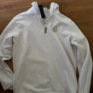 Vit half zip sweatshirt i bomull S - Vit sweatshirt i storlek small med half zip och hög krage. Tröjan har ett broderat märke på bröstet och är tillverkad i mjuk bomull. Perfekt för lager-på-lager och chill dagar. Långärmad och enkel design som funkar till det mesta.