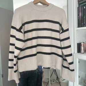 Randig beige och svart tröja H&M - Säljer en beige stickad tröja från H&M med svarta horisontella ränder. Tröjan har breda ribbade muddar, rund hals och en loose fit. Materialet är en mix av polyester, akryl, polyamid och ull.