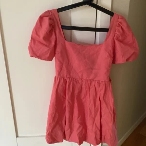 Rosa kort puffärmad klänning med knyt - Superfin rosa kortklänning med puffärmar och fyrkantig urringning. Klänningen har en söt knytdetalj i ryggen och en puffig kjol nertill. Perfekt för sommarens alla tillfällen och ger en riktigt söt vibe.