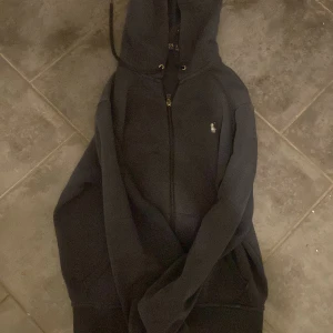 Mörk blå hoodie från Polo Ralph Lauren - Mörk blå hoodie från Polo Ralph Lauren med dragkedja och klassisk broderad logga på bröstet. Tröjan har huva med snörning och två fickor framtill. Perfekt för en chill och stilren look.