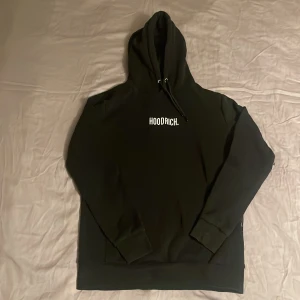 Svart hoodie från Hoodrich - Svart hoodie från Hoodrich med vit logga på bröstet. Klassisk modell med huva och snörning, samt långa ärmar och ribbade muddar. Perfekt för dig som gillar streetwear och vill ha en enkel men snygg look. Ena plastbiten till snöret är sönder, förutom det är den i väldigt bra skick. Dm för bild.