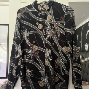 Svart mönstrad skjorta från H&M - Snygg svart skjorta från H&M i storlek 36 med ett detaljerat paisleymönster i vitt, beige och rött. Skjortan har långa ärmar, klassisk krage och knäppning framtill. Perfekt för dig som gillar unika prints och vill sticka ut lite extra.