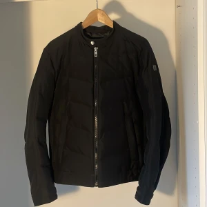 Svart pufferjacka från Boss Orange - Svart pufferjacka från Boss Orange med stilren design och raka linjer. Jackan har hög krage, dragkedja framtill och diskret logga på ärmen. Perfekt för dig som gillar en clean look och vill hålla värmen under kyliga dagar. Nypris ligger på omkring 3-3500.