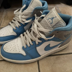 Nike Air Jordan 1 Mid UNC blå/vit - Nike Air Jordan 1 Mid i klassisk UNC-färg. Skorna har en snygg kombination av ljusblått och vitt skinn med gråa detaljer och vit sula. Ikonisk Jordan-logga på sidan och tungan, samt snörning framtill. Perfekta för dig som gillar streetwear och sneakers med retrovibe.