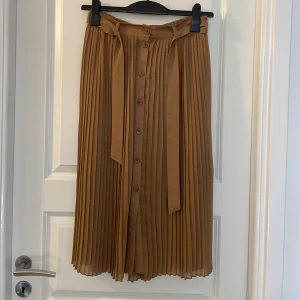 Plisserad beige midikjol från Monki - Säljer en snygg plisserad beige kjol från Monki i storlek S. Kjolen har knappar framtill, ett matchande bälte i midjan och är tillverkad i ett lätt, flowy material. Perfekt för dig som gillar en stilren och trendig look.