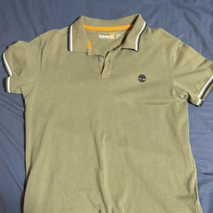 Olivgrön pikétröja från Timberland - Snygg olivgrön pikétröja från Timberland med klassisk krage, knappar och broderad logga på bröstet. Tröjan har korta ärmar och vita ränder på krage och ärmslut. Perfekt för en avslappnad och stilren look.