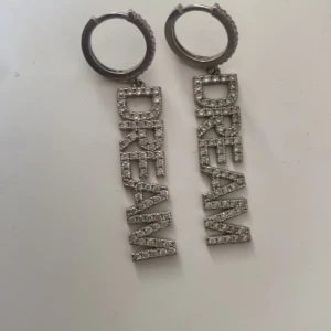 Ioaku dream örhängen  - Säljer ett par statement örhängen i silverfärg med hängande texten 'DREAM' täckt av glittrande stenar. Bokstäverna är stora och i blockstil, vilket ger en cool och trendig vibe. Perfekt för dig som gillar att sticka ut med accessoarer. Från ioaku som är helt nickelfria.