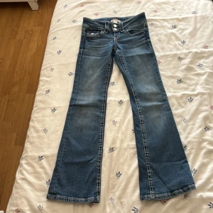 Blå bootcut jeans från Gina Tricot - Snygga blå jeans från Gina Tricot i storlek 134. Modellen har bootcut-ben, klassiska fem fickor och tydliga kontrastsömmar. Jeansen är i denim med en ljus tvätt och har dubbla knappar framtill för extra detalj.